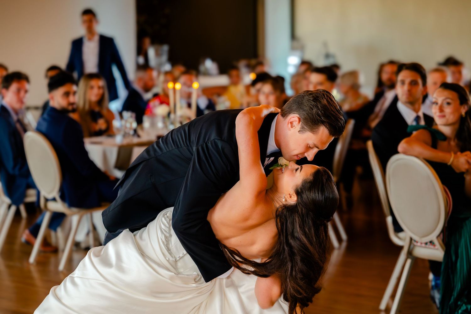 Groom dips bride for a romantic kiss on the dance floor surrounded by wedding reception guests.