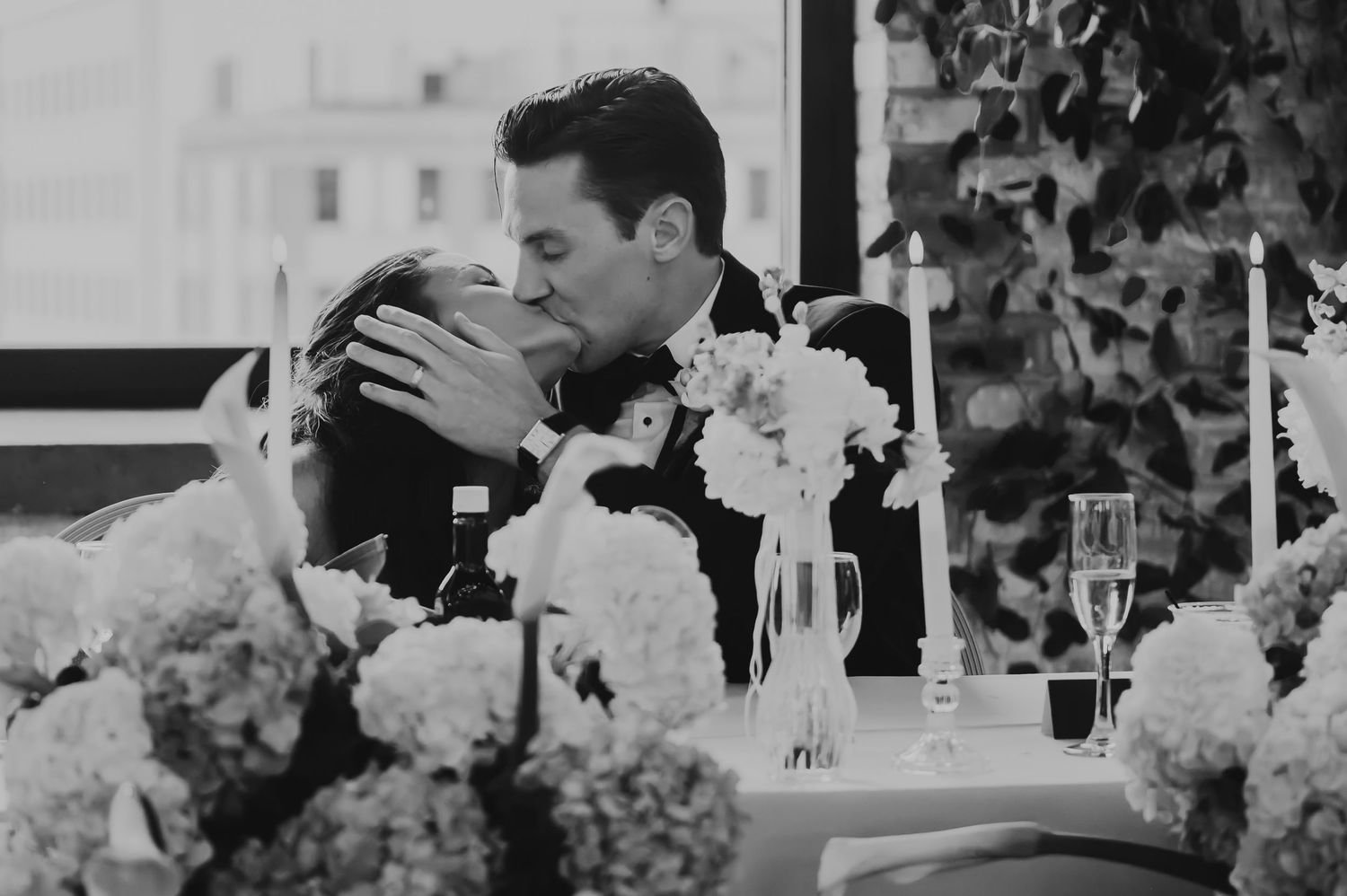 Black and white photo of a couple sharing a romantic kiss at their wedding reception table surrounded by white floral arrangements.