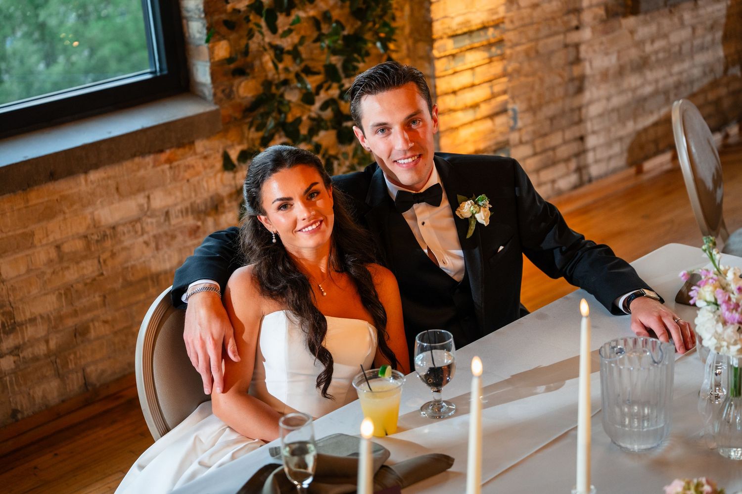 Smiling bride and groom sit together at their wedding reception table with candles and drinks nearby.