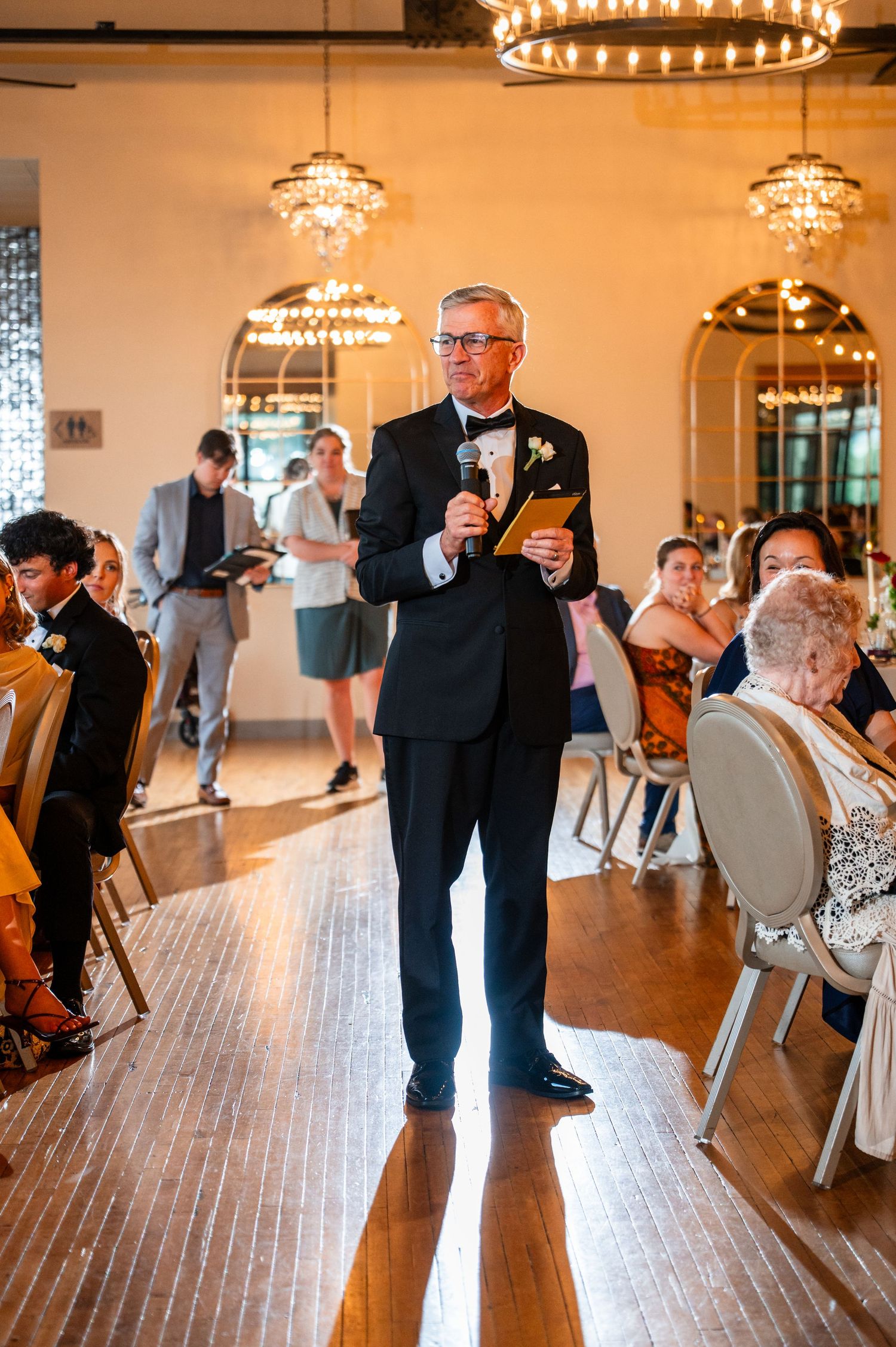 Man in black tuxedo delivers a heartfelt wedding speech with a microphone in a chandelier-lit venue.