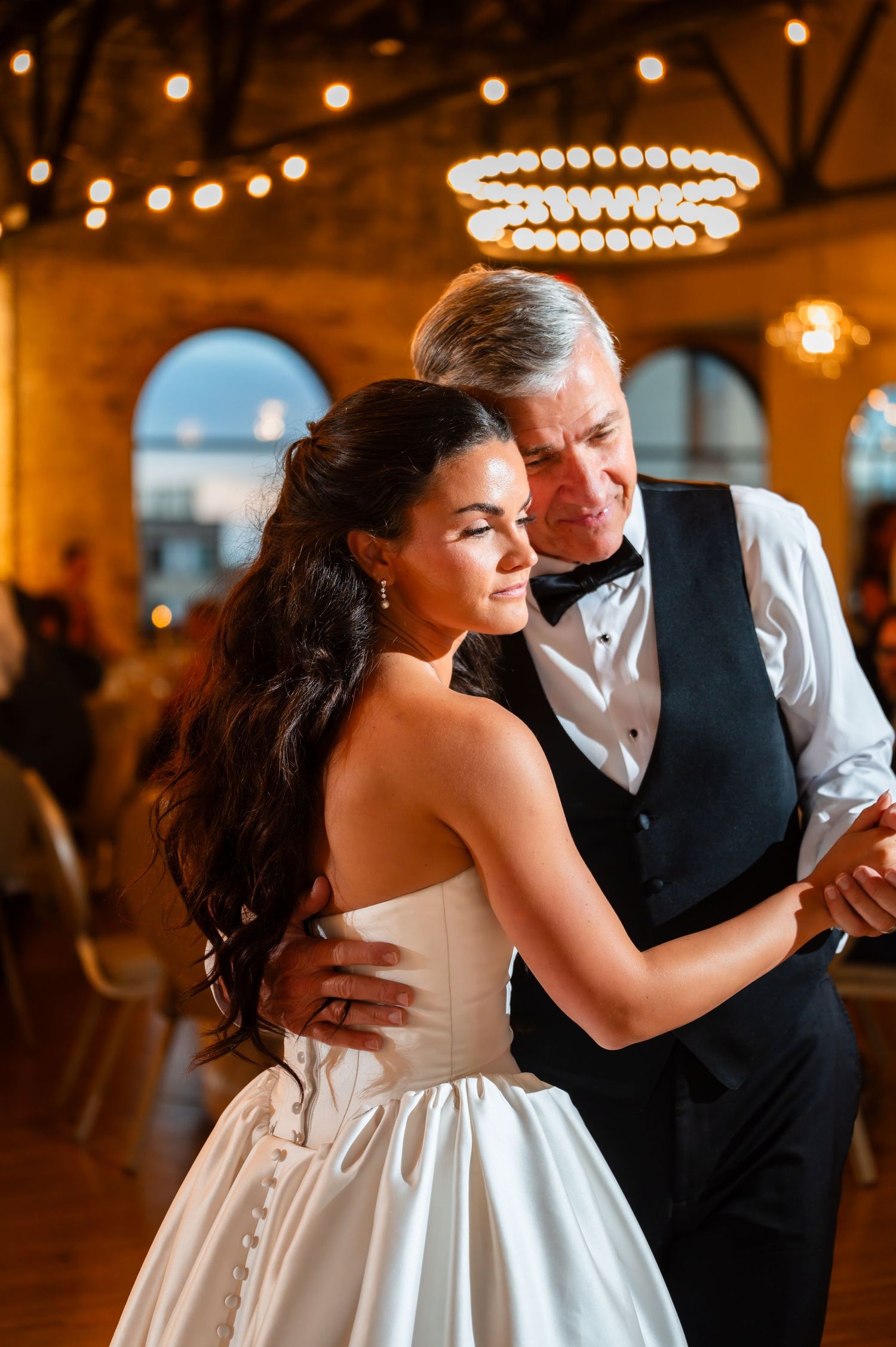 Bride shares an emotional father-daughter dance under warm string lights at a rustic wedding reception.
