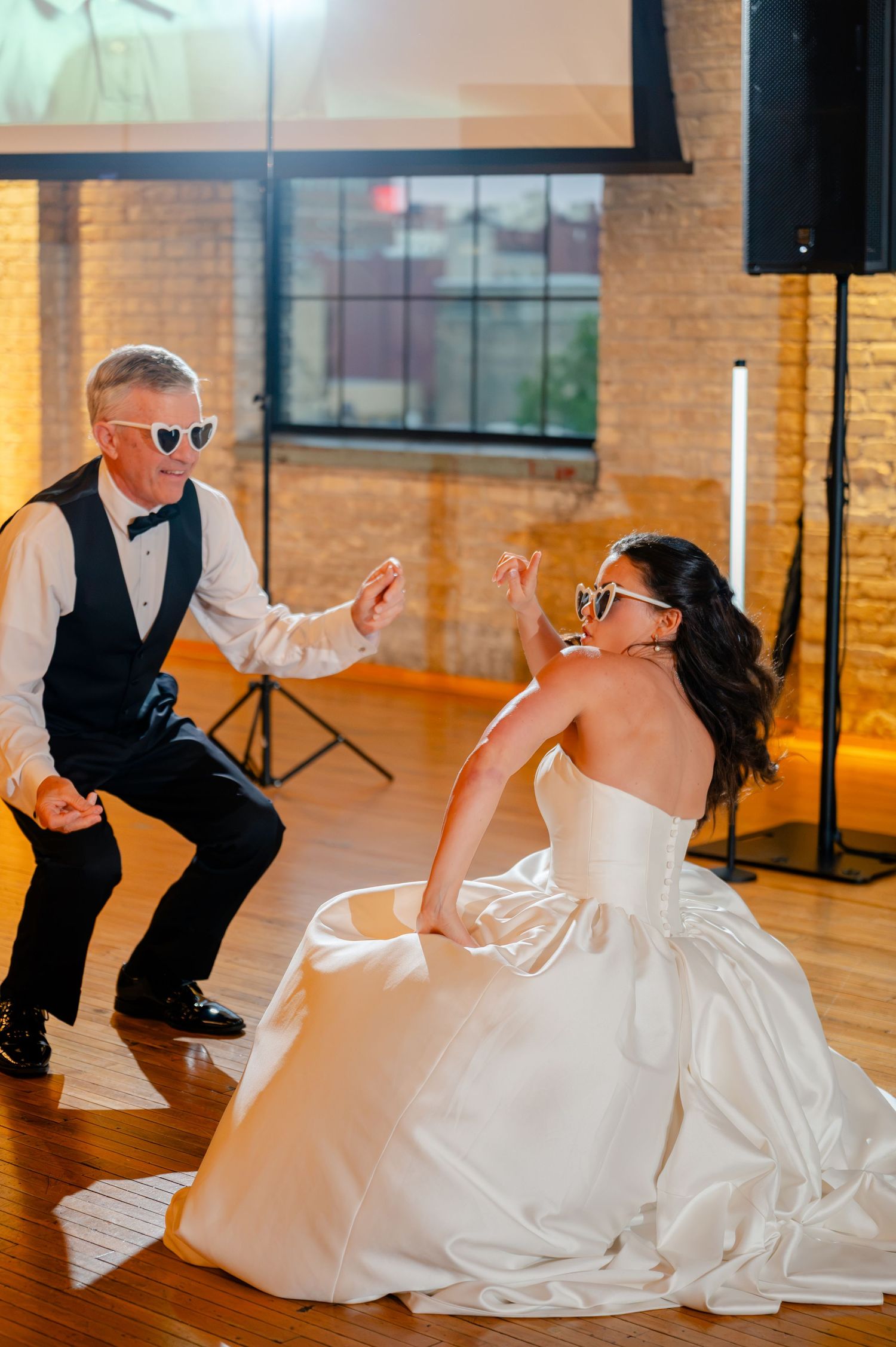 Bride and older gentleman dance together wearing fun heart-shaped sunglasses at wedding reception.