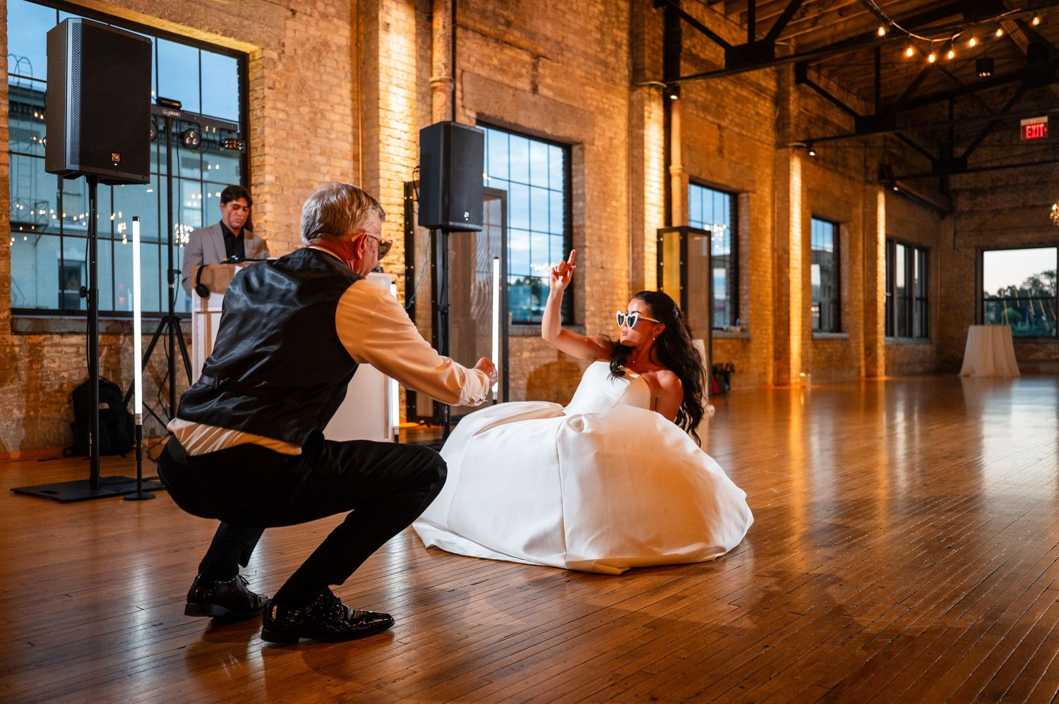 Bride in white ball gown dances with guest on warm-lit hardwood floor in rustic brick reception venue.