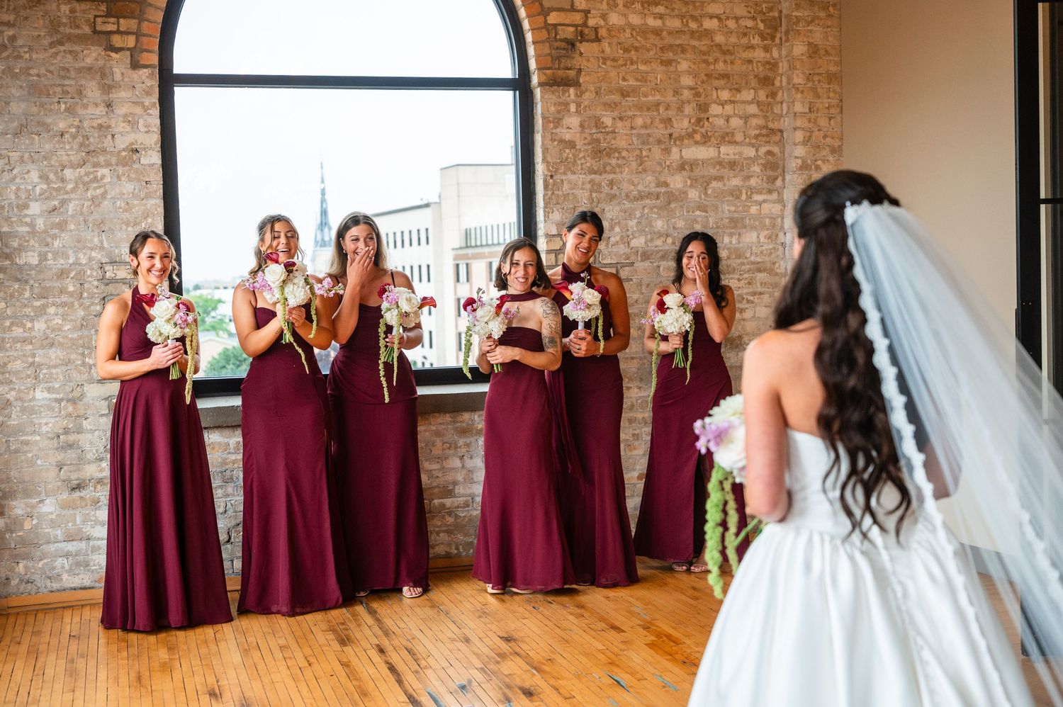 Bridesmaids in burgundy gowns react emotionally during a first look reveal with the bride in a white wedding dress.