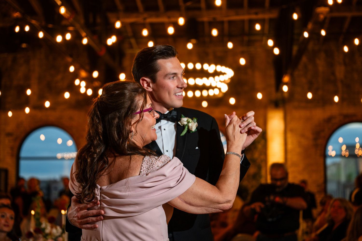 Groom dances joyfully with his mother under glowing string lights at a warm wedding reception.