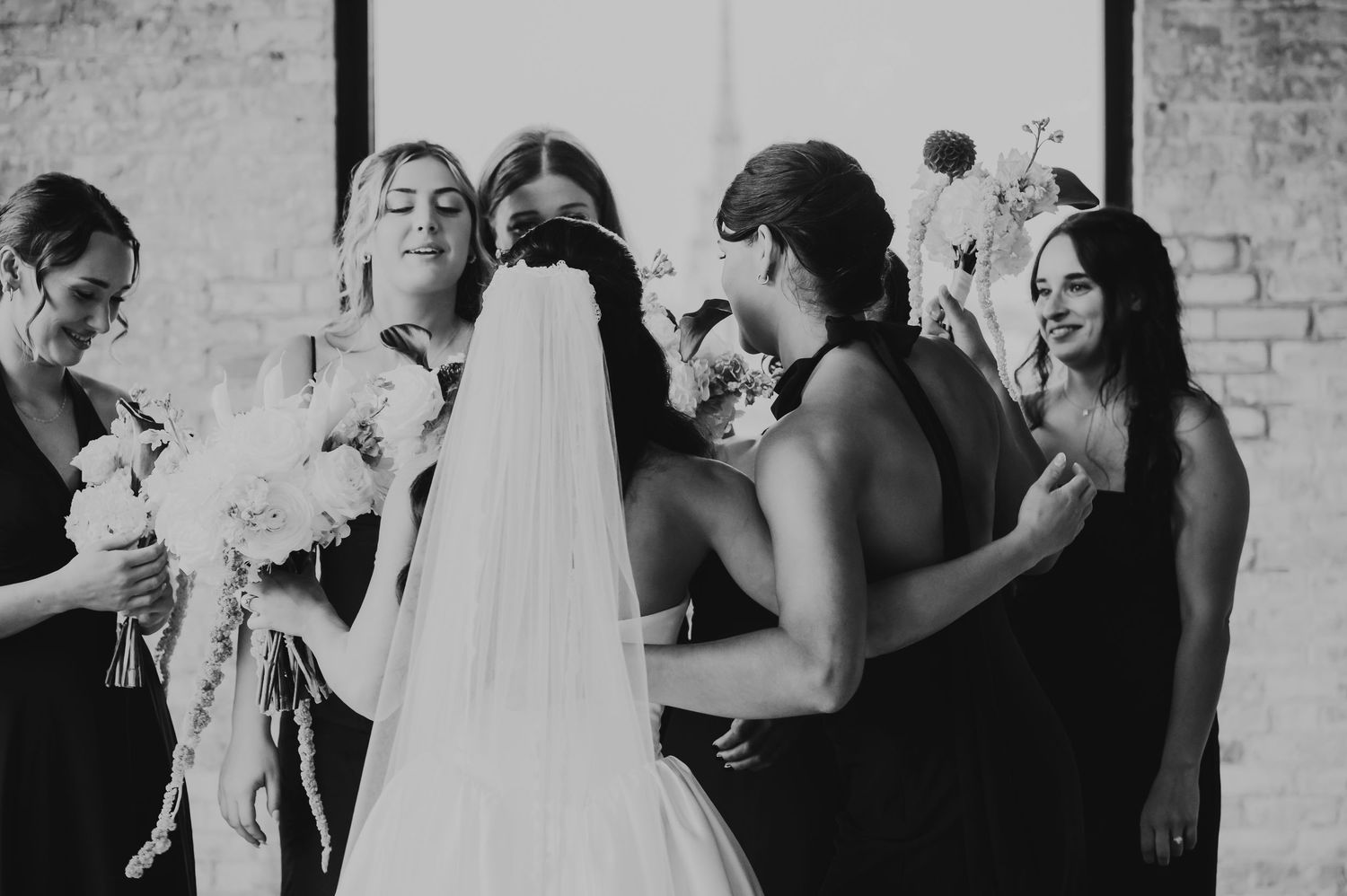 Bridesmaids in black dresses surround a bride in white gown and veil, sharing a joyful moment before the wedding ceremony.