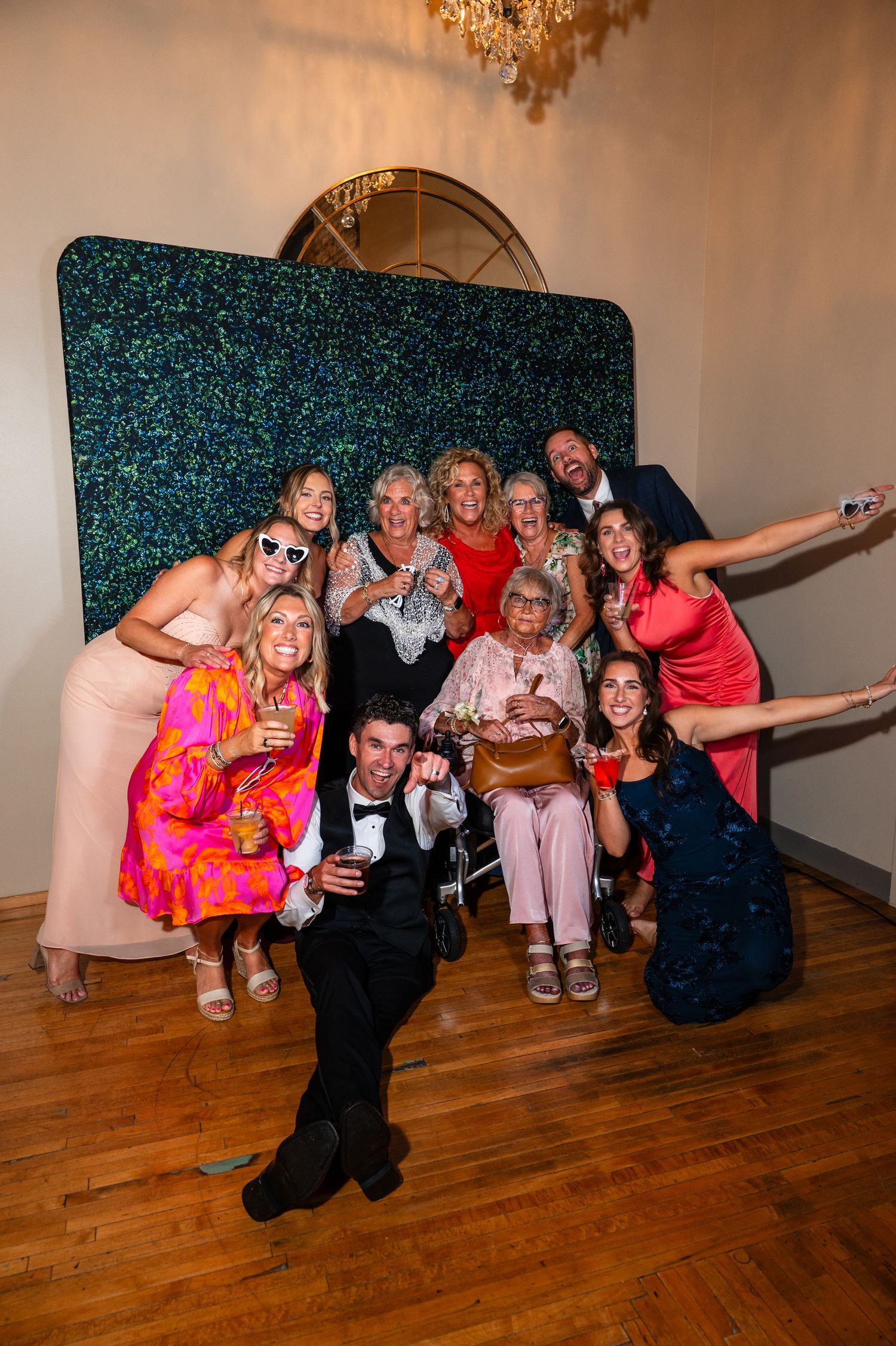 Joyful wedding guests pose together at photo booth with green sequin backdrop during reception celebration.