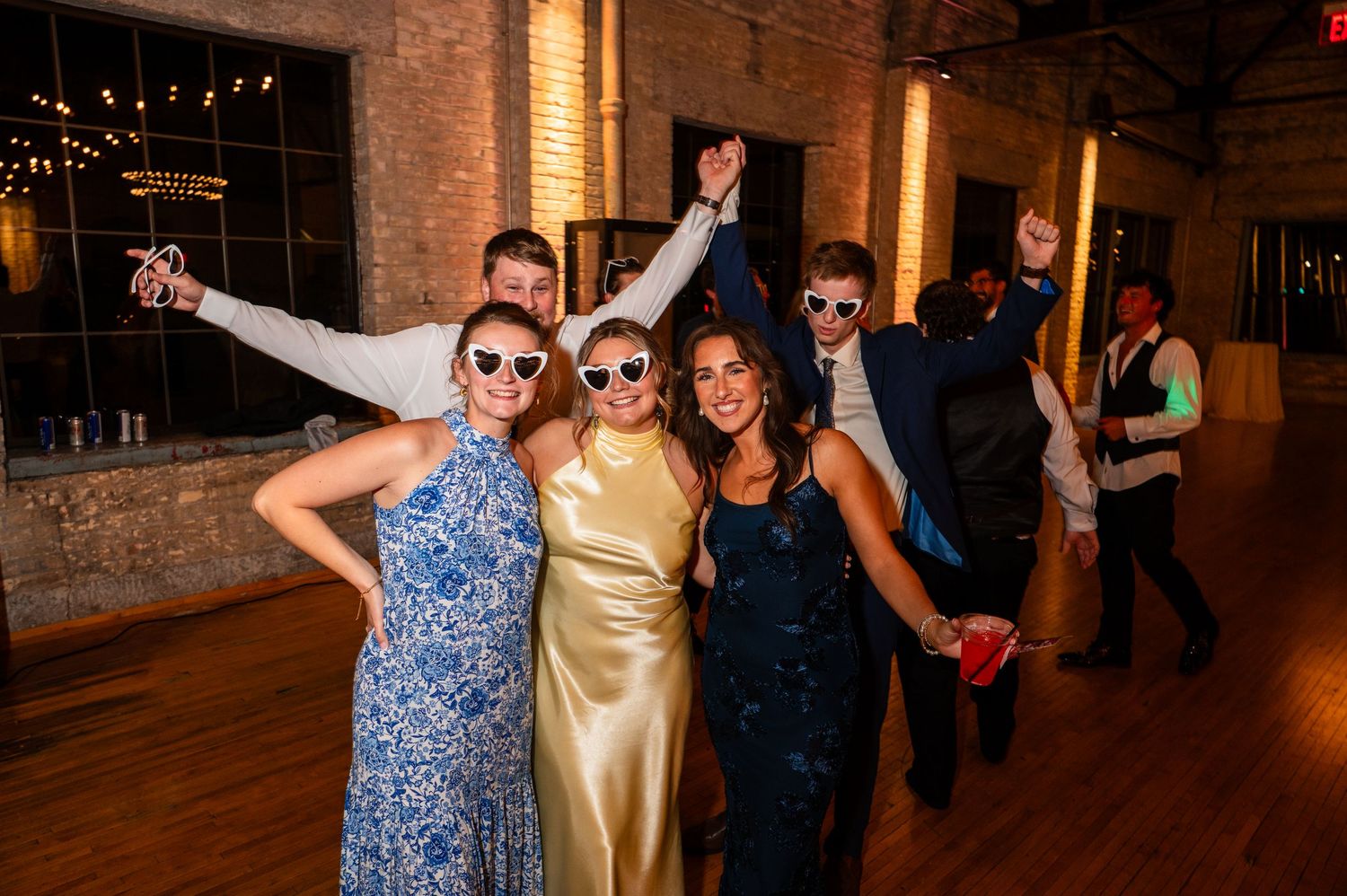 Smiling wedding guests wearing novelty sunglasses dance together on the floor at evening reception.