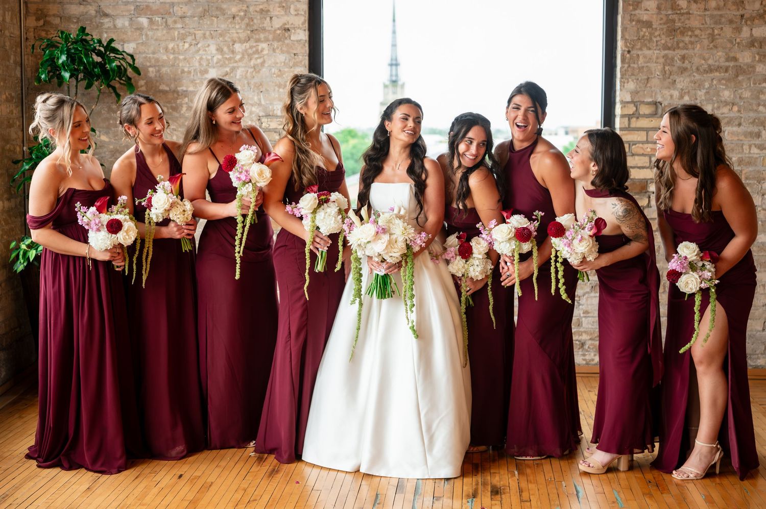 Bride in white gown with bridesmaids in burgundy dresses holding floral bouquets at an indoor wedding venue.