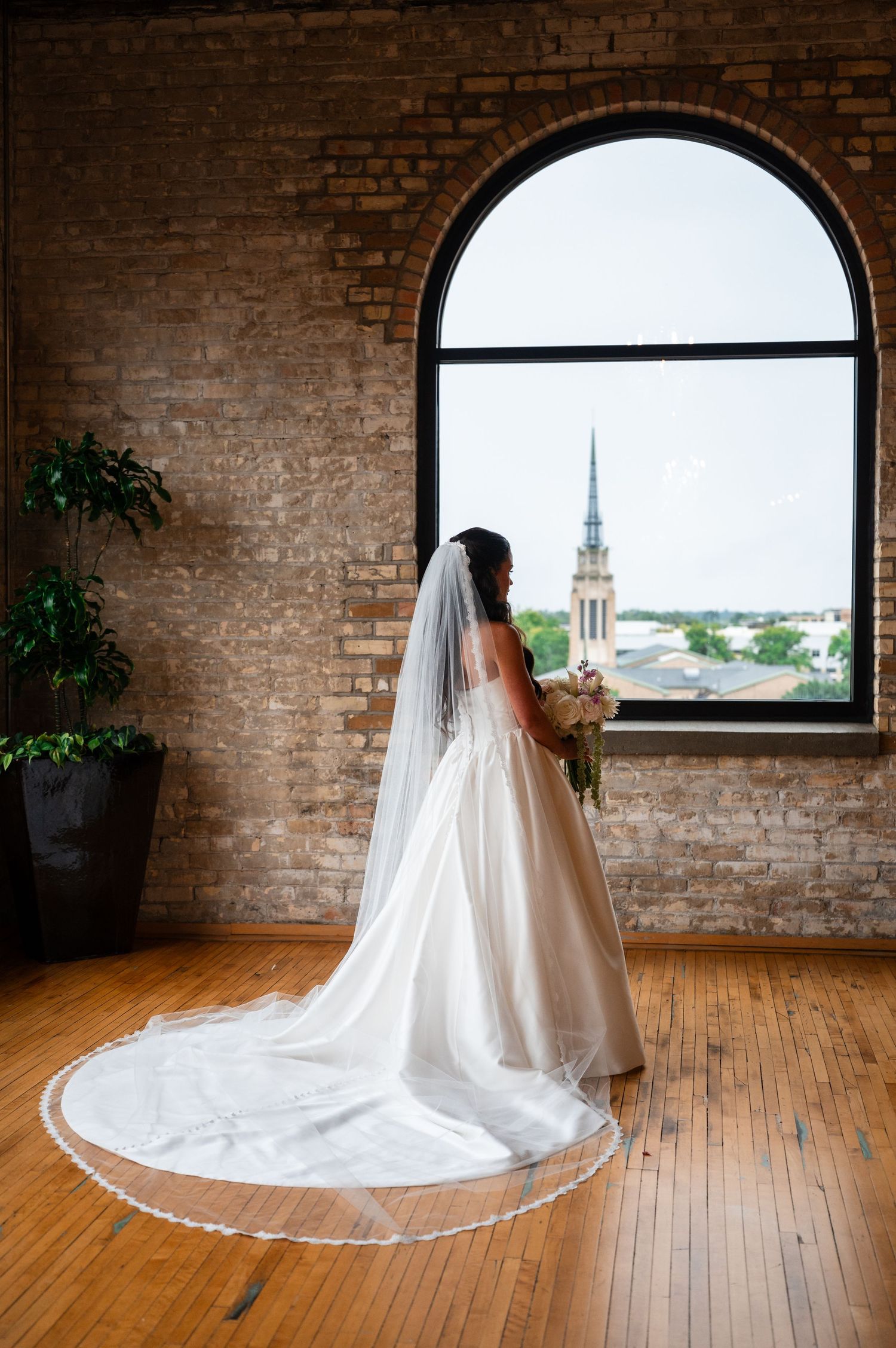 Bride in a white gown with long cathedral veil stands before arched window with city views in brick venue.