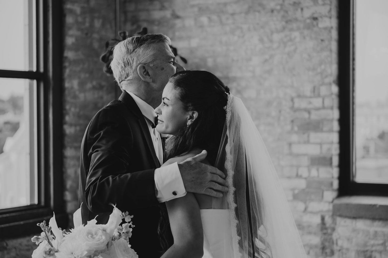 Black and white photo of a bride in a veil sharing a tender embrace with an older man in a suit at a wedding.