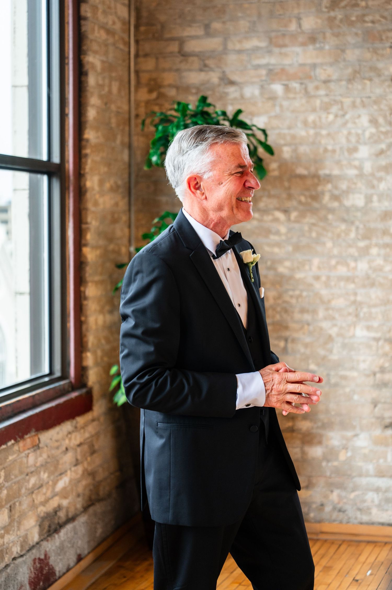 Smiling older man in black tuxedo with boutonniere stands by brick wall and window at wedding.