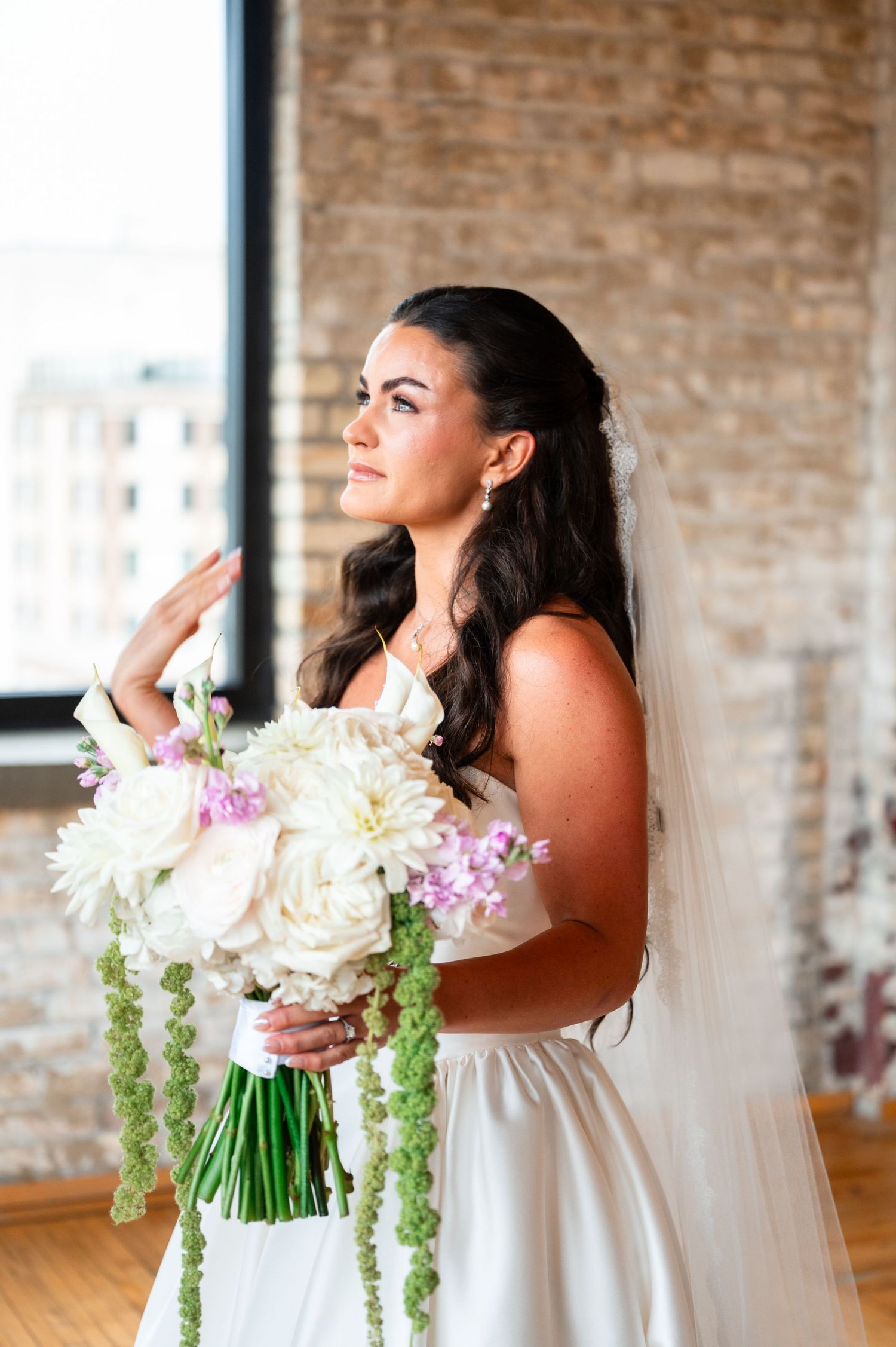 Beautiful bride holding white floral bouquet with greenery wearing veil by brick wall window.