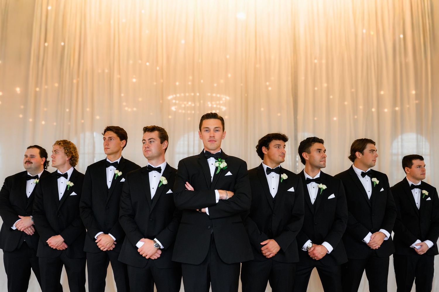Groom and groomsmen in black tuxedos with bow ties pose elegantly in front of a white draped backdrop.