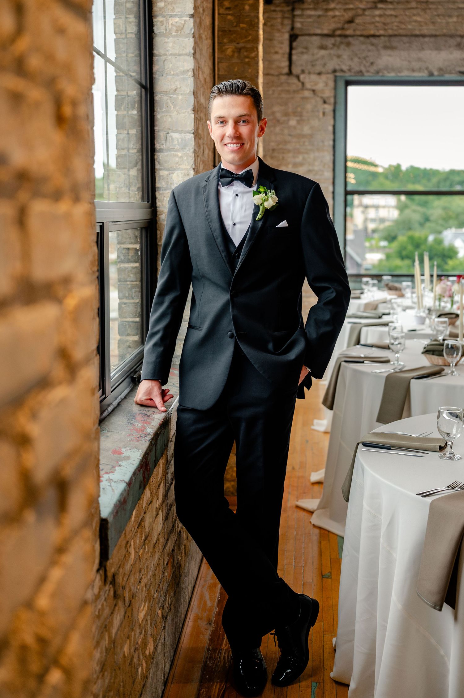 Groom in black tuxedo with bow tie and boutonniere leaning against brick wall in elegant wedding venue.