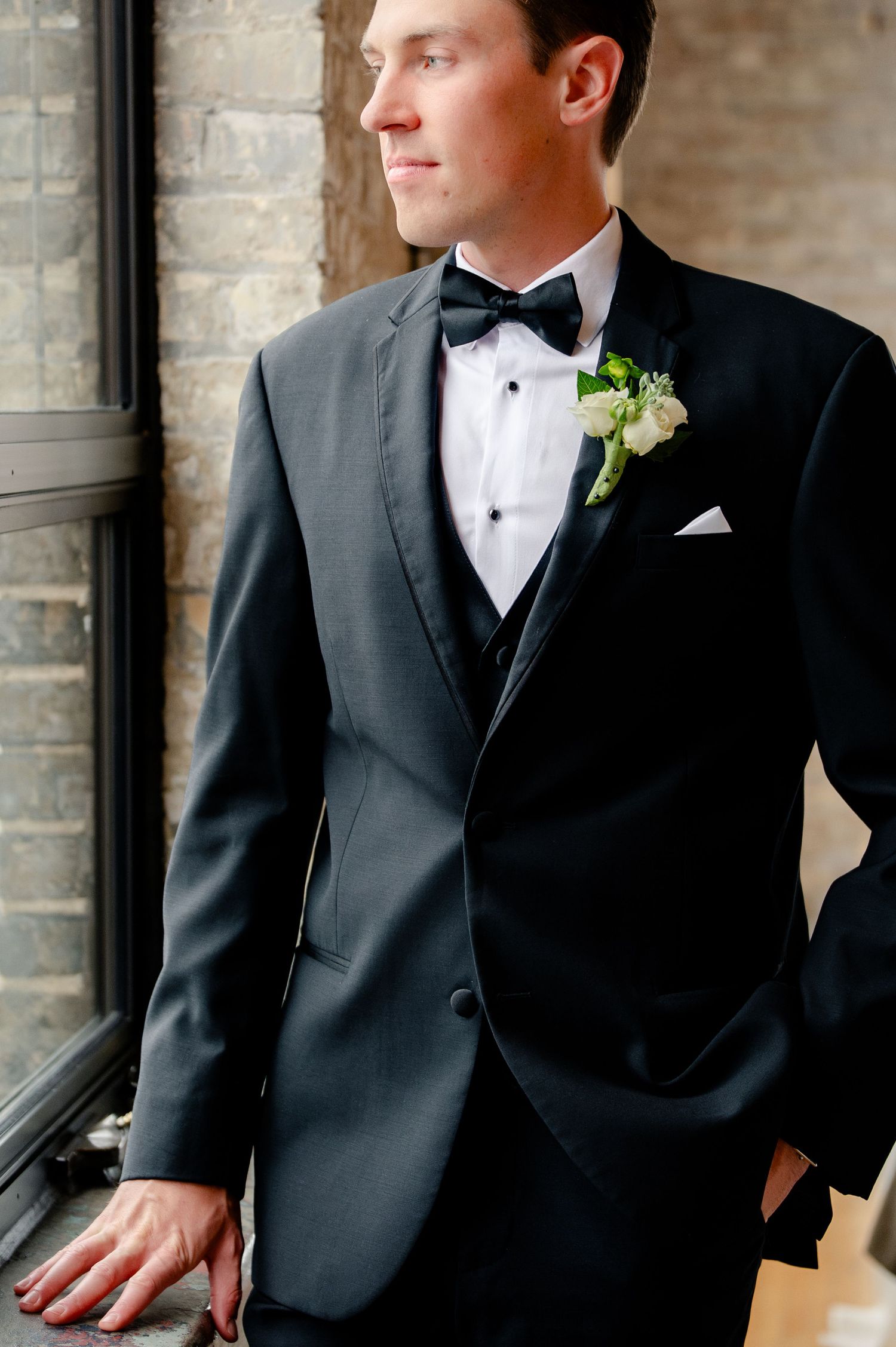 Groom in black tuxedo with bow tie and white boutonniere poses elegantly near a window on his wedding day.