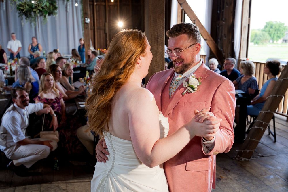 Bride and groom in a pink suit share their first dance during a rustic wedding reception at The Centennial Barn.