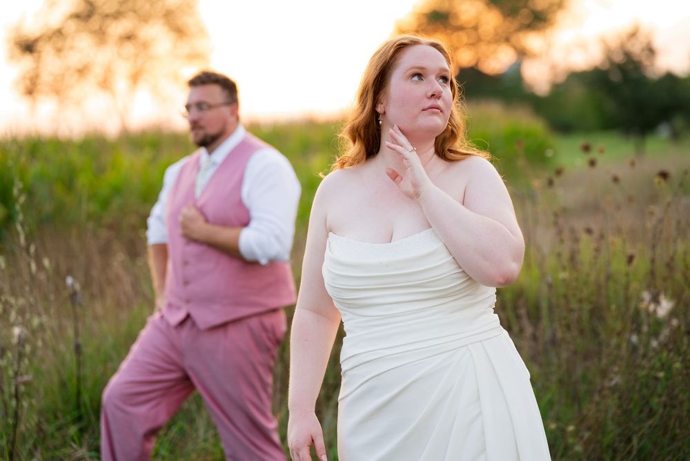 A bride and groom pose during golden-hour portraits in a late-summer field. She is featured in front, he is in the background.