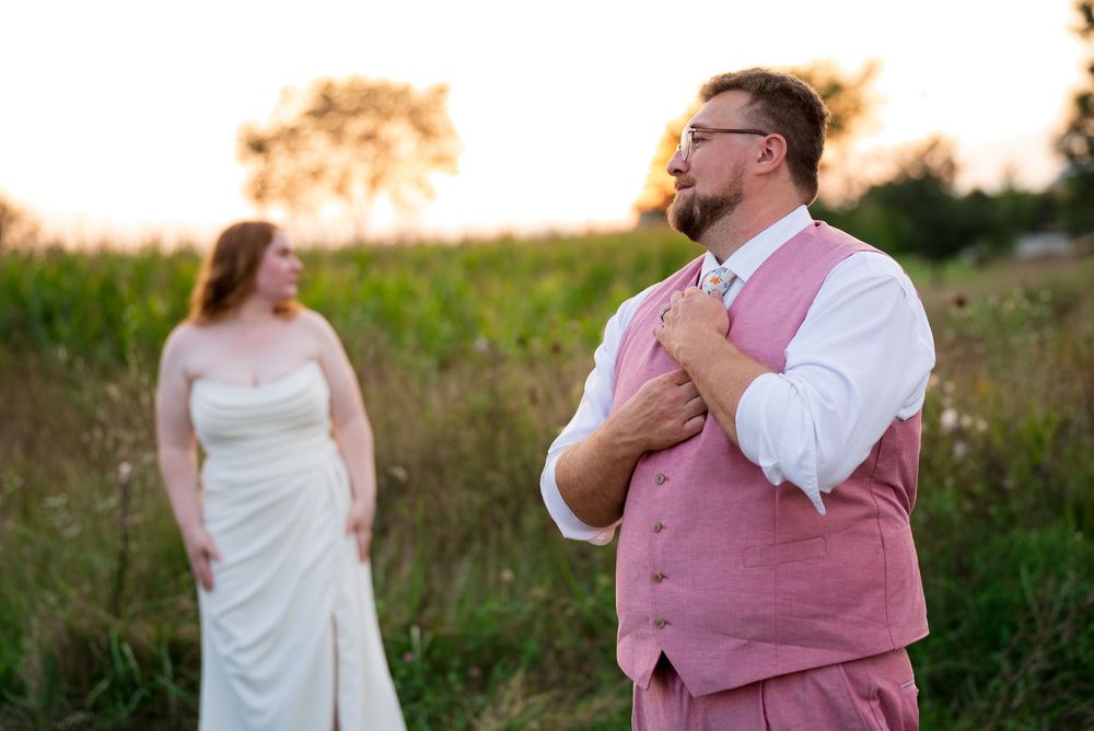 A bride and groom pose during golden-hour portraits in a late-summer field. He is featured in front, she is in the background.