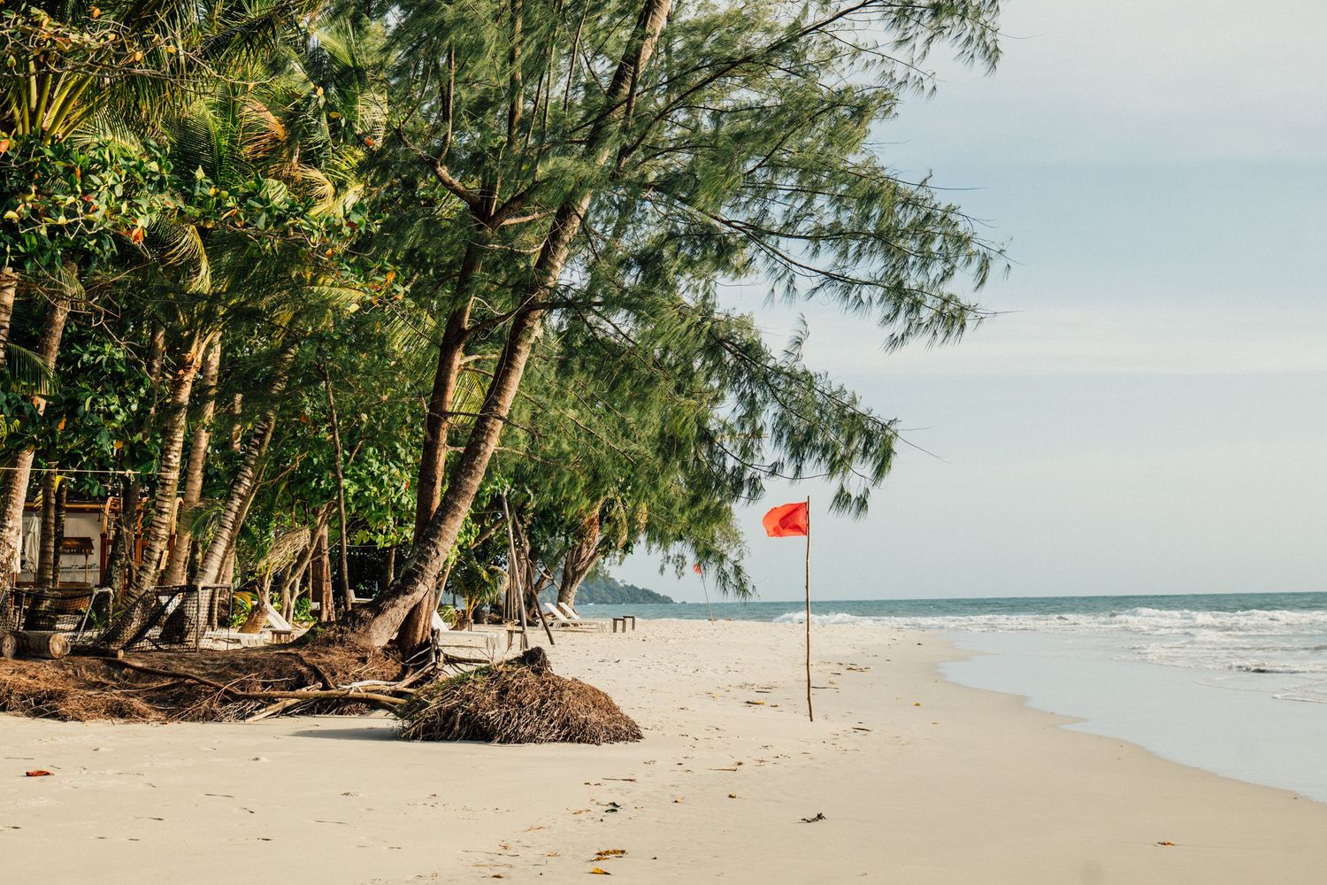 Tropical beach with tall palm trees, white sand, and a red flag near calm ocean waves under a partly cloudy sky.
