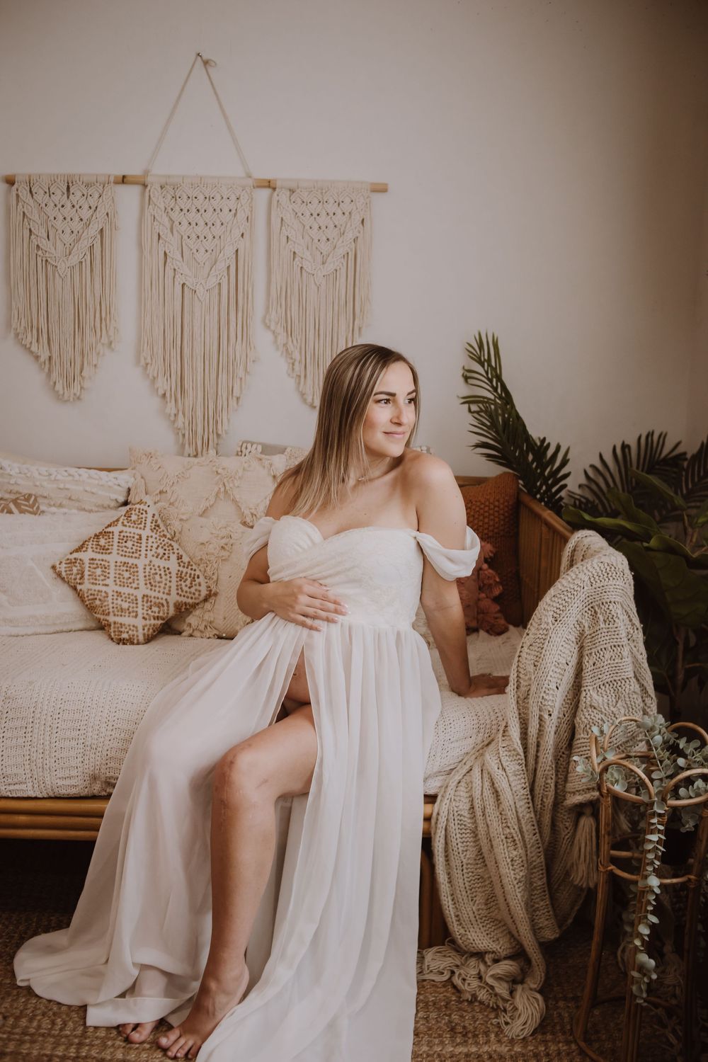 Pregnant woman in white off-shoulder gown sitting in boho bedroom with macrame wall decor.