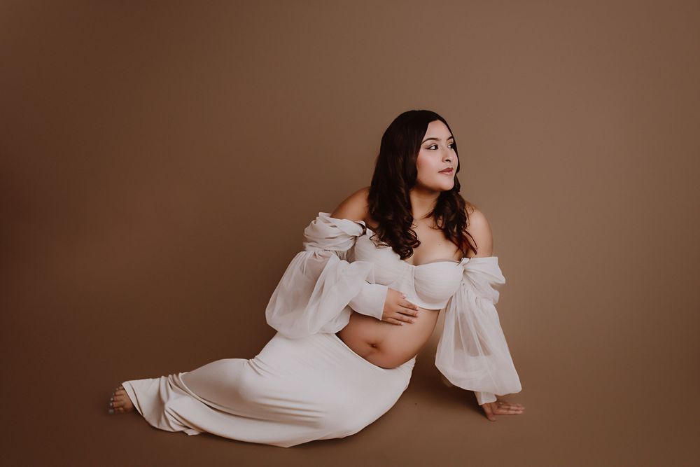 Pregnant woman in white off-shoulder gown sitting gracefully against brown backdrop in elegant maternity photo session.