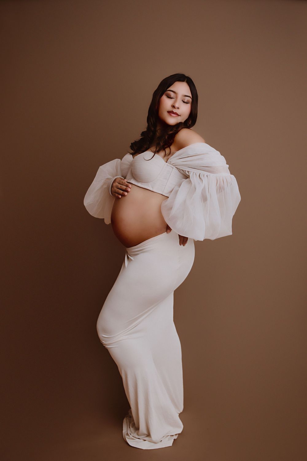 Pregnant woman in white off-shoulder gown cradling baby bump against brown studio background.