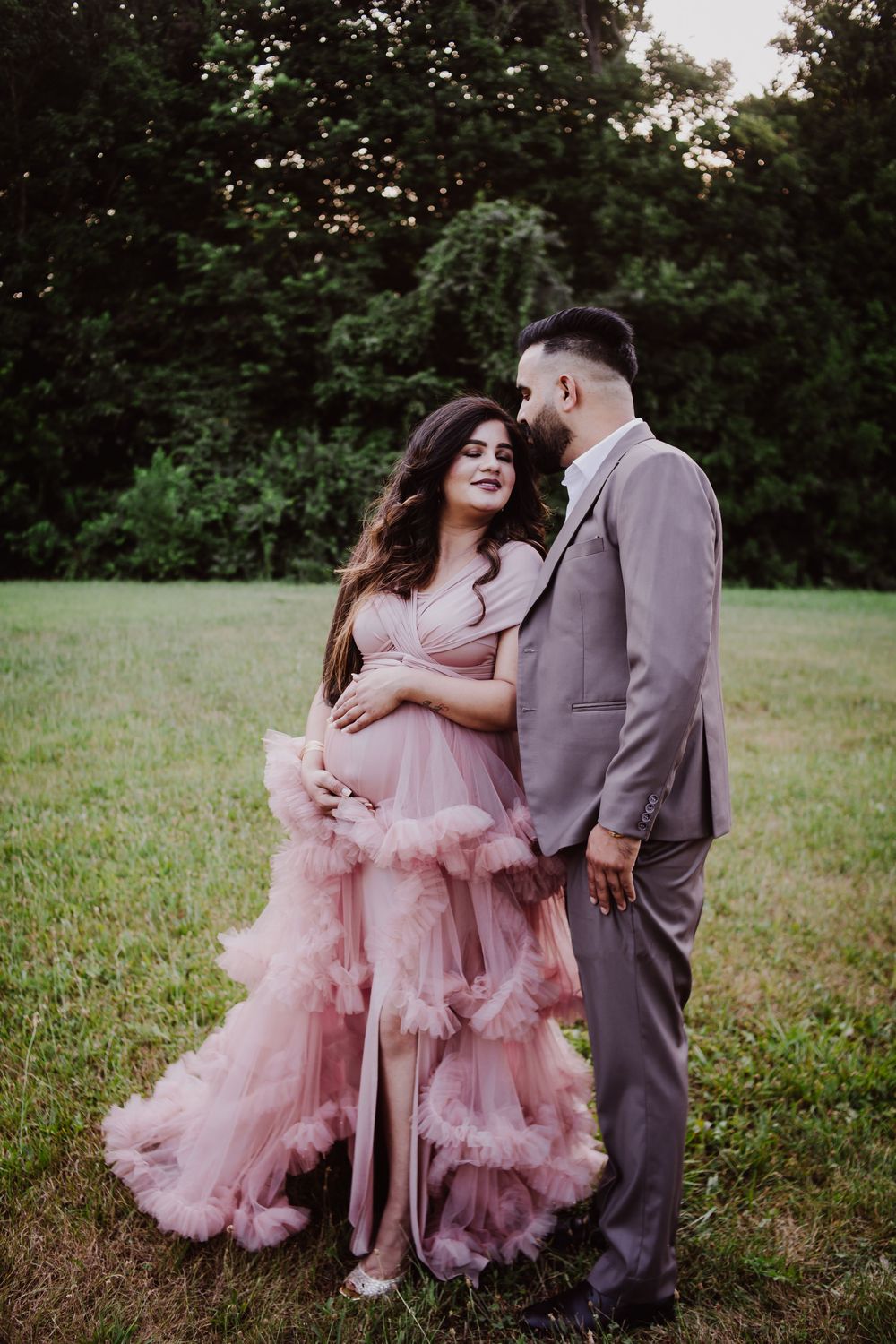 Couple in maternity photoshoot, woman in pink ruffle gown, man in gray suit, standing in green field.