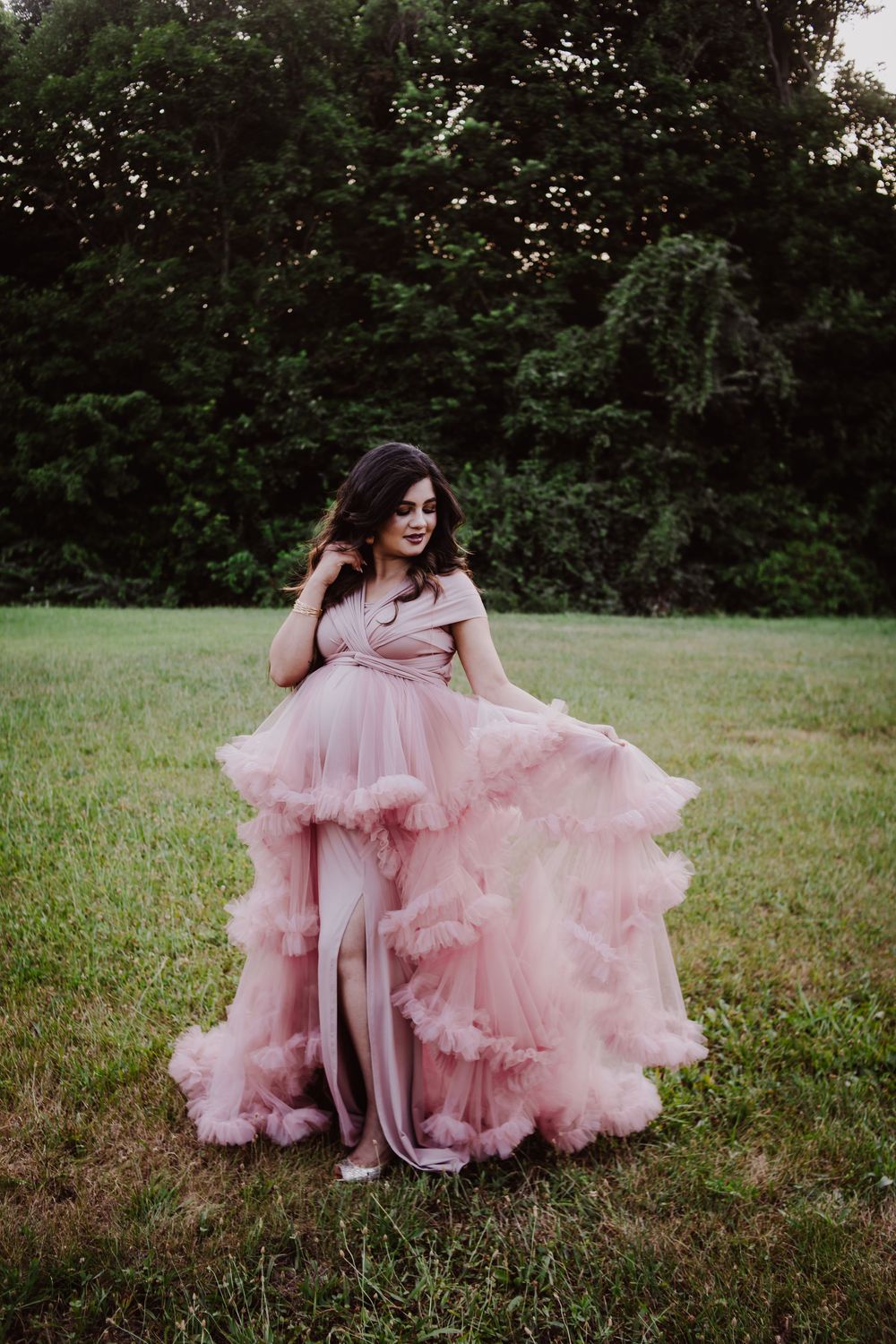 A woman in a stunning pink ruffled tulle gown poses gracefully outdoors on green grass with lush trees behind her.
