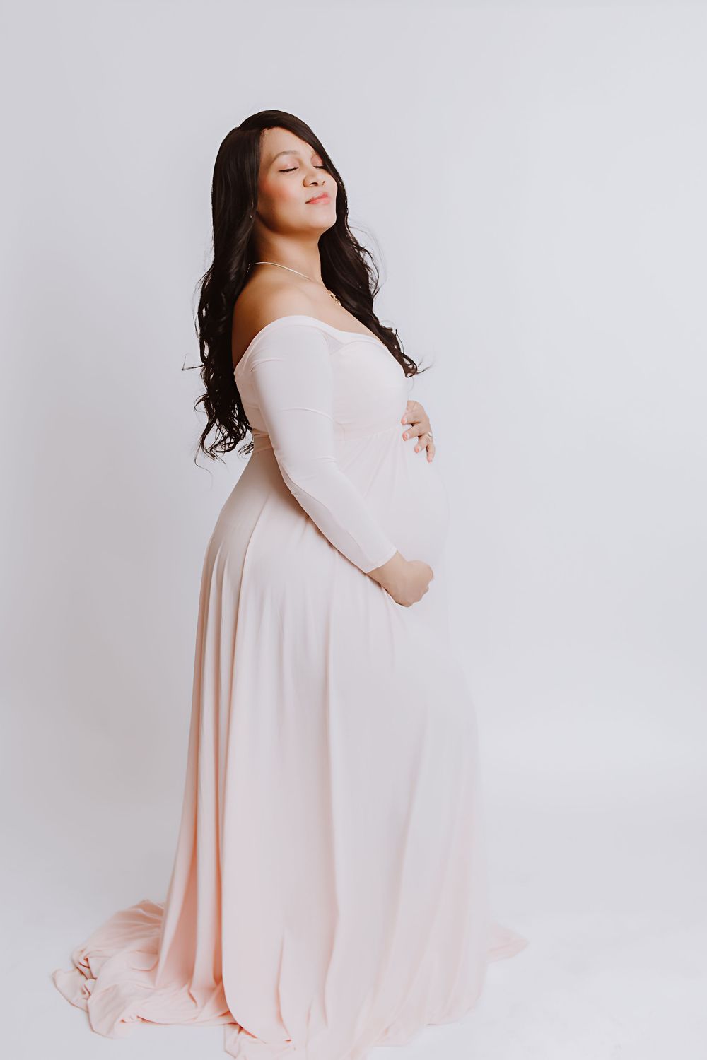 Pregnant woman in elegant blush off-shoulder gown cradling baby bump, eyes closed, studio maternity portrait.