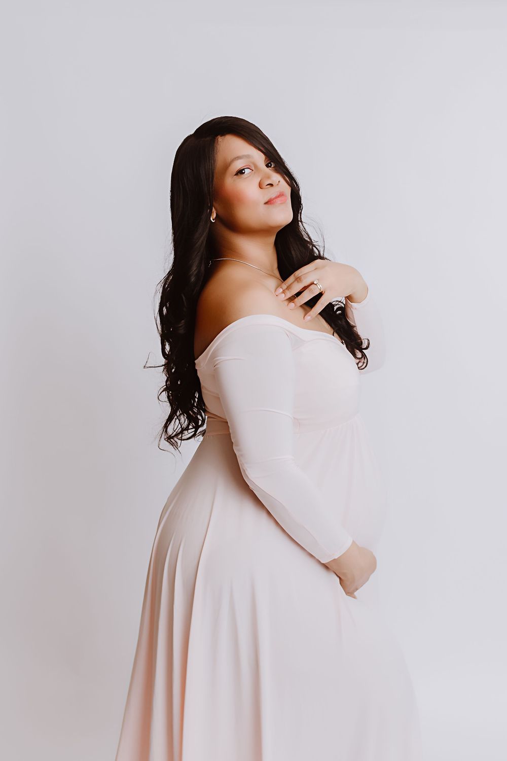 Pregnant woman in off-shoulder blush pink maternity gown posing gracefully against white background.