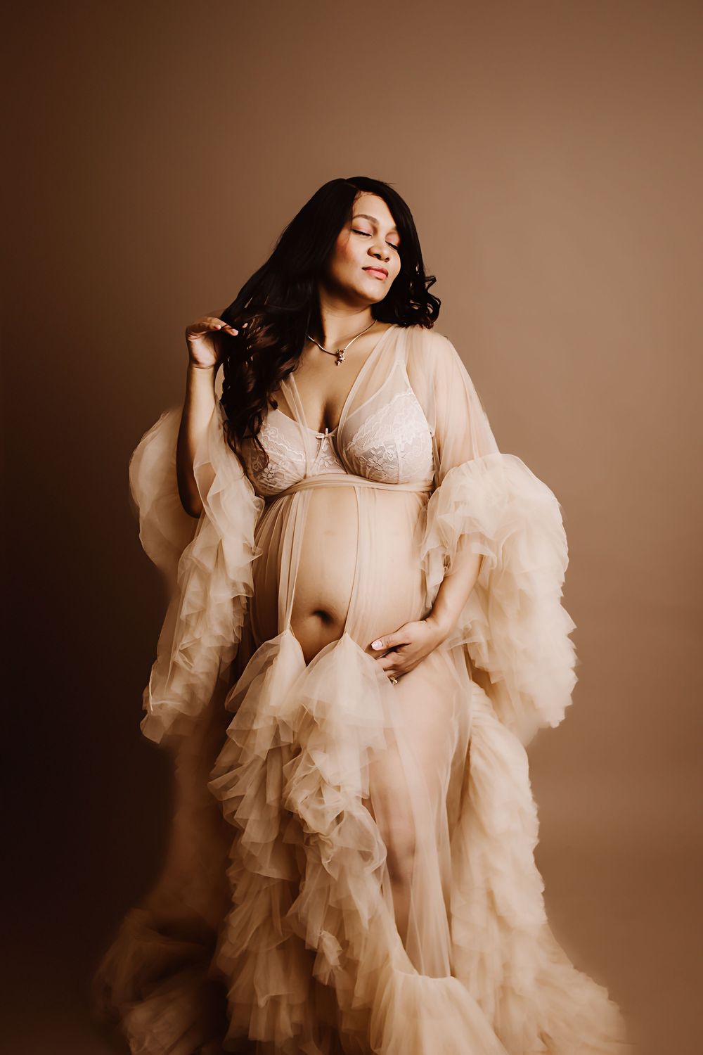 Pregnant woman in sheer ruffled maternity gown poses gracefully against warm brown studio background.