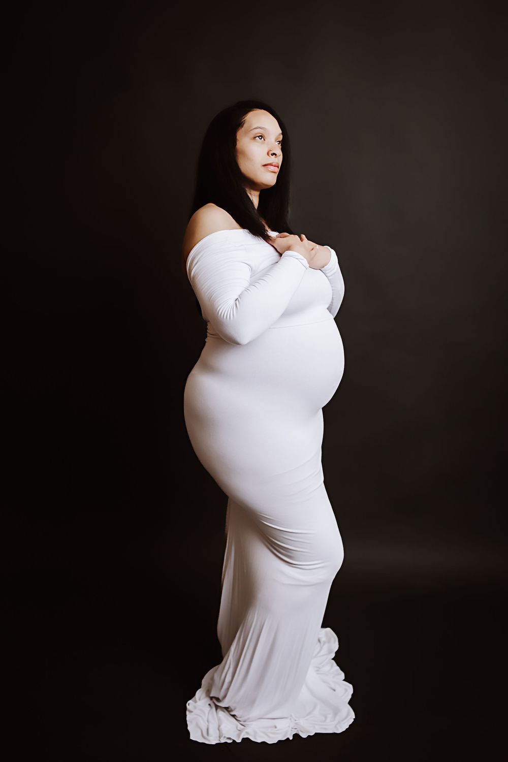 Pregnant woman in white off-shoulder gown poses elegantly against dark background, cradling her baby bump.
