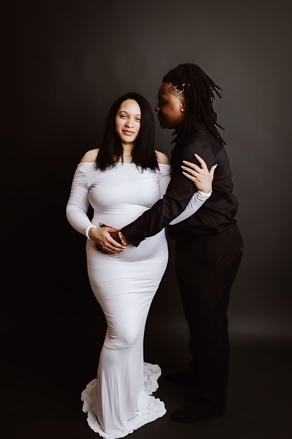 A couple poses together, woman in white off-shoulder gown embraced by partner in black outfit against dark studio background.