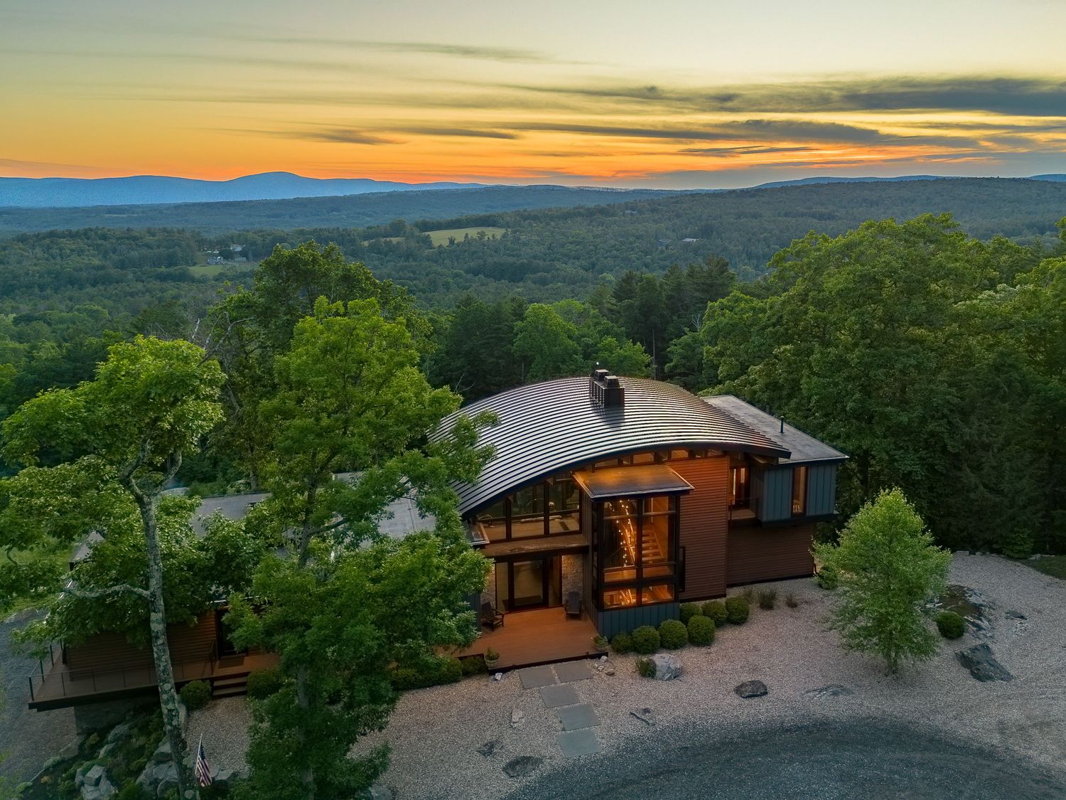 Stunning aerial view of a modern hilltop home surrounded by trees with panoramic mountain views at sunset.