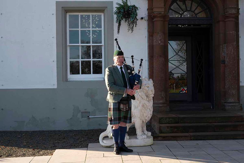 A Scottish bagpiper in traditional tartan kilt performs outside an elegant manor house entrance with a stone lion statue.