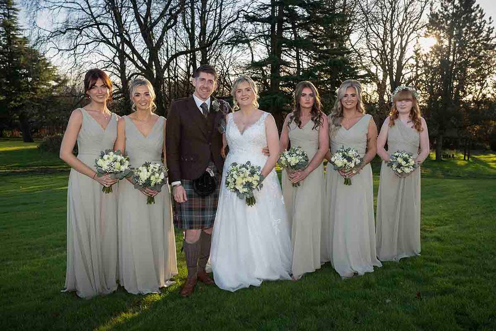 Groom in tartan kilt stands with bride in white gown and five bridesmaids in champagne dresses holding white bouquets outdoors.