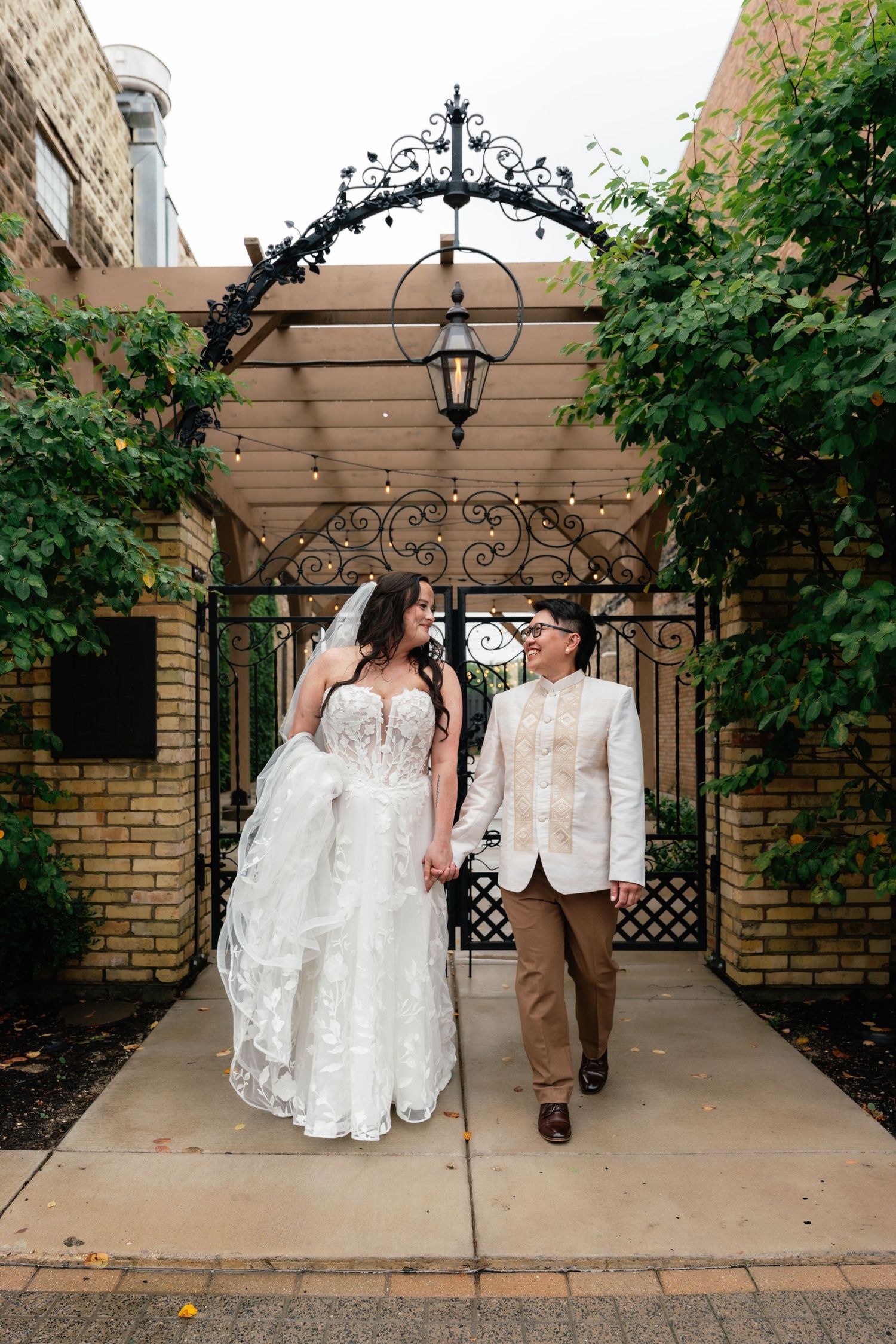 Two brides share a first kiss at their wedding, standing before a brick wall with a neon sign.