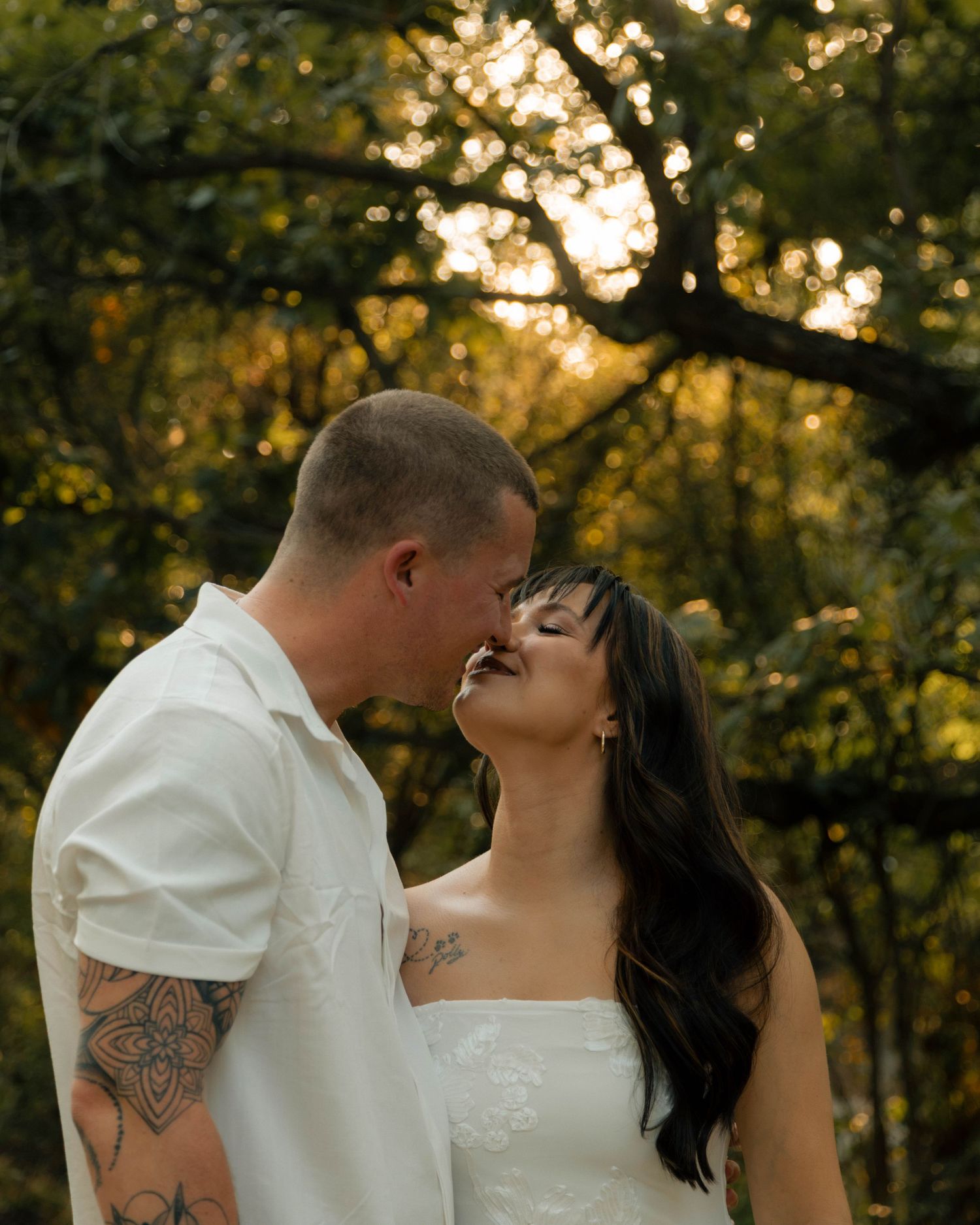Couple sharing a romantic almost-kiss outdoors at golden hour, man with tattoos in white shirt, woman in white dress.