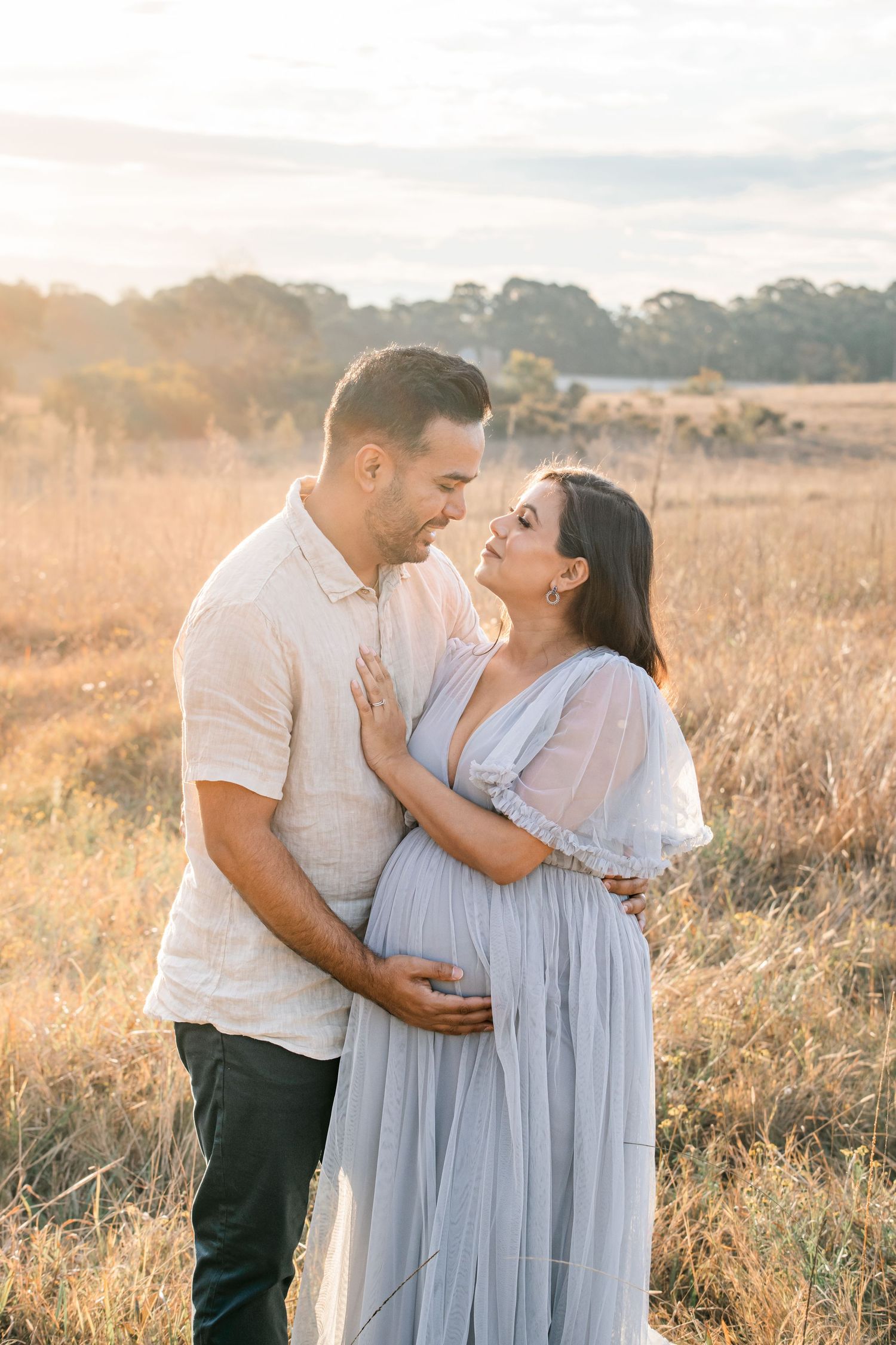 Couple embracing in a golden field at sunset in Rouse Hill, woman in grey cradling her bump.