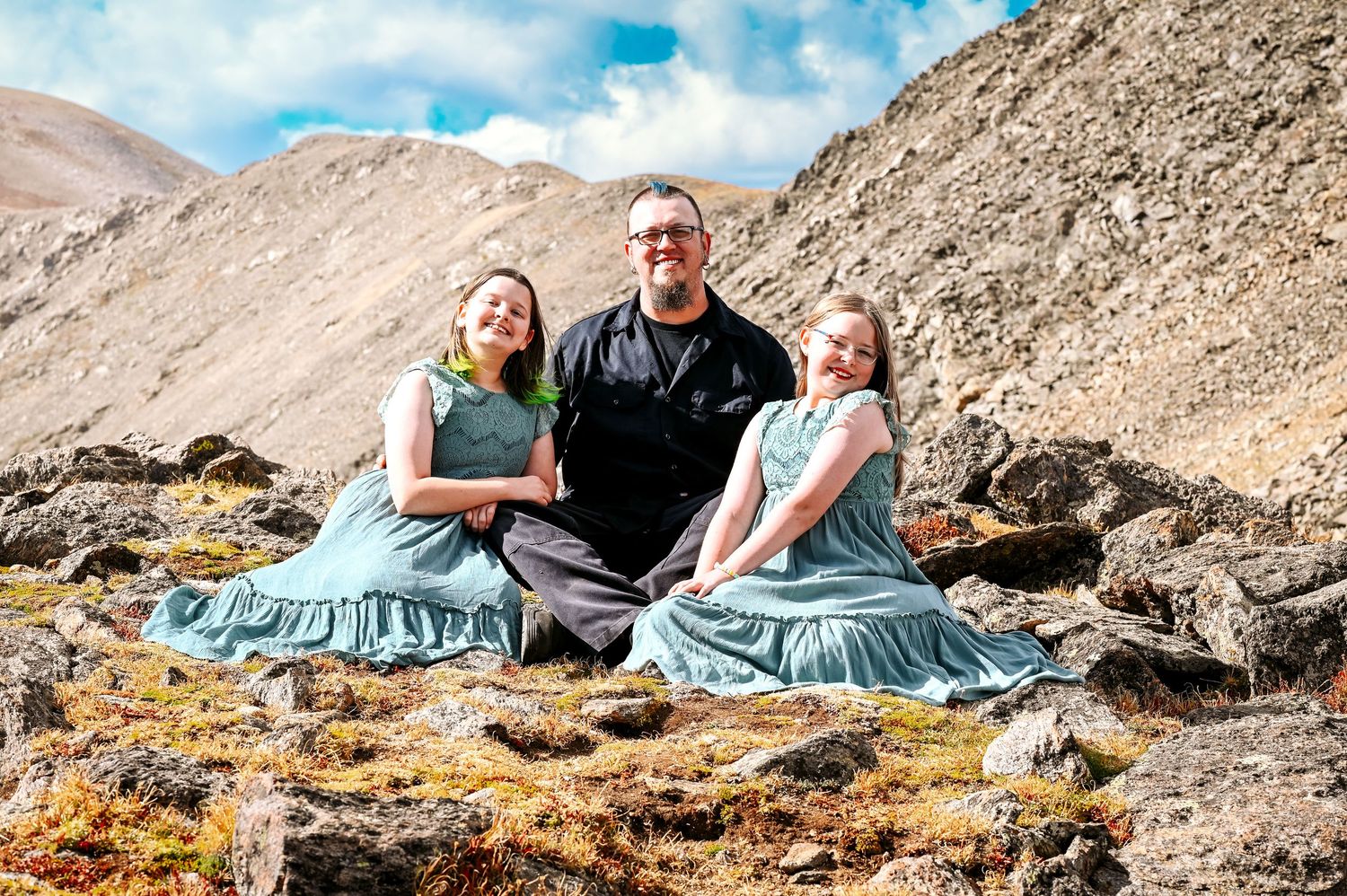 Man and two girls in teal dresses posing on rocky mountain terrain under a partly cloudy sky.
