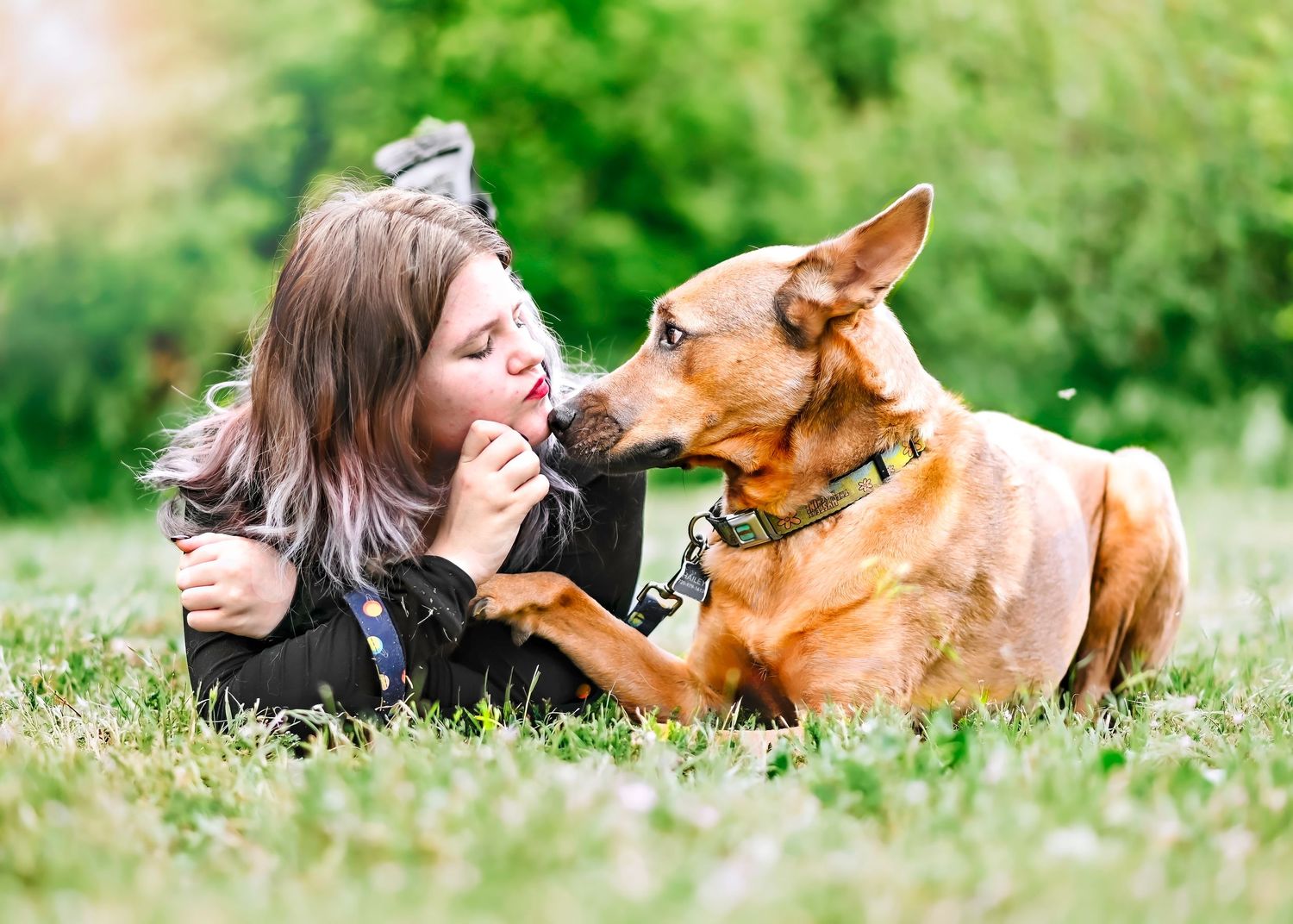 A young woman lying on grass shares a tender moment with her brown dog wearing a green collar outdoors.