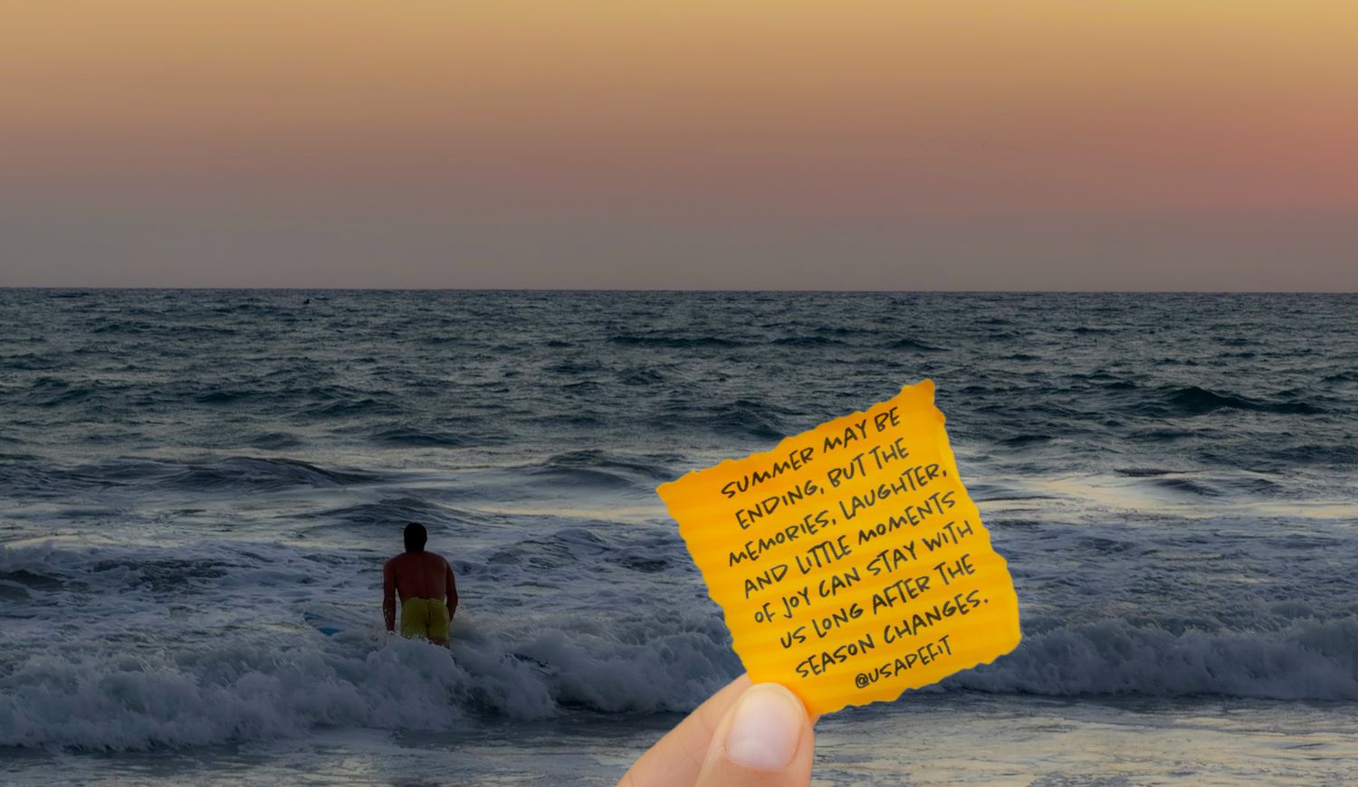 Person standing in ocean waves at sunset holding a yellow sticky note with inspirational quote, warm sky backdrop.