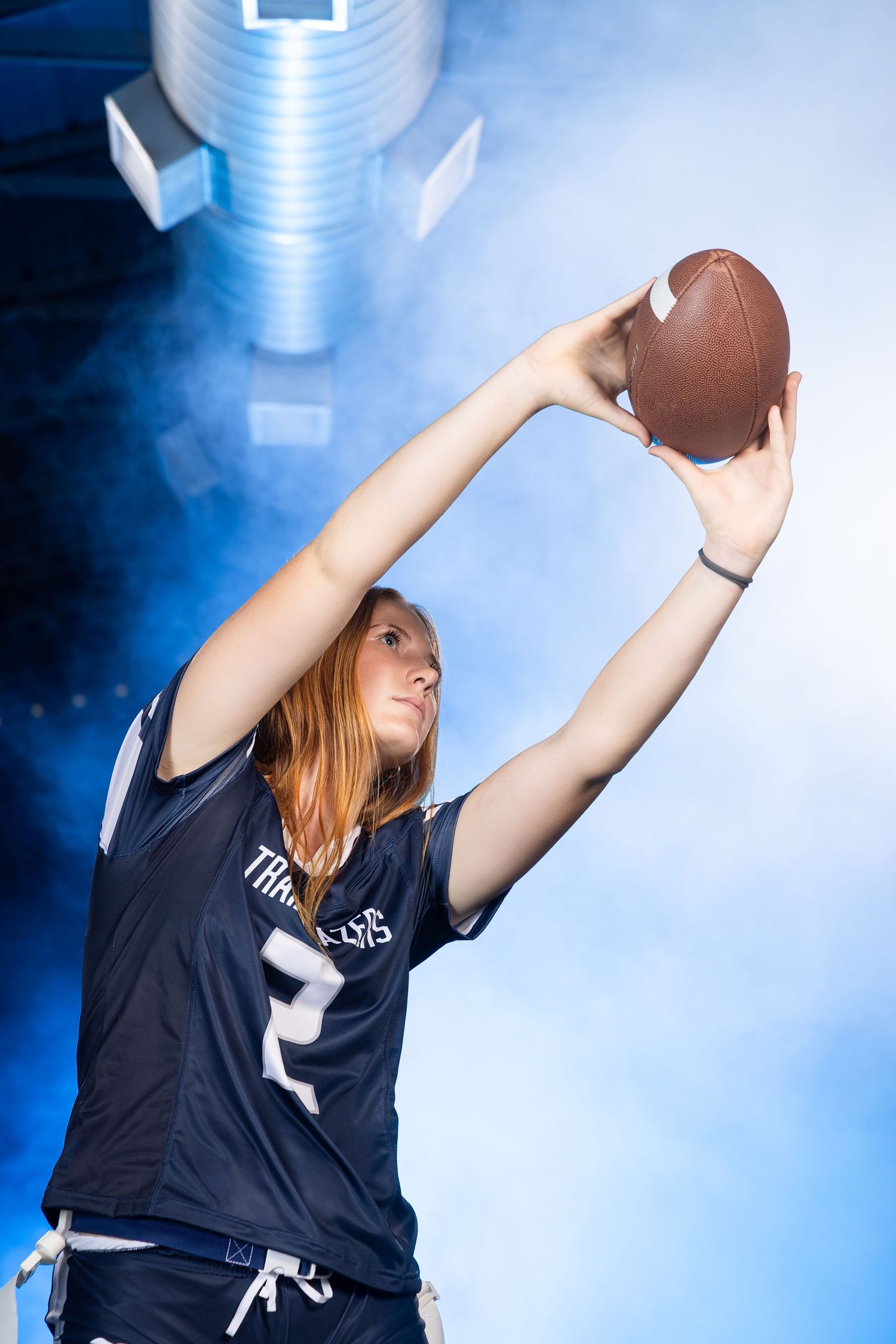Young woman in football jersey holds ball overhead in dramatic blue smoky stadium lighting.