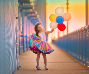 A young girl in a colorful rainbow dress holds a bouquet of balloons on a bridge at sunset.