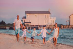A happy family of five holds hands walking along a beach at sunset near a waterfront home.