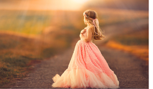 A little girl in a flowing pink ball gown walks down a sunlit country road at golden hour.