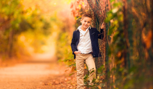 A smiling boy in a navy jacket leans against a tree on a colorful autumn forest path.