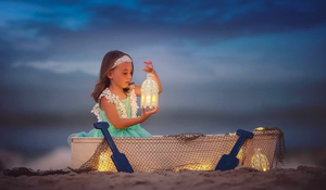 A little girl in a teal dress holds a glowing lantern while sitting in a basket on the beach at dusk.