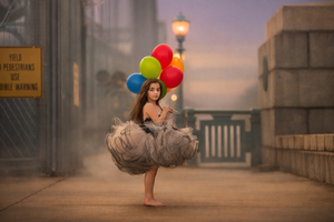 A barefoot girl in a gray tutu dress holds colorful balloons on a misty urban bridge at dusk.